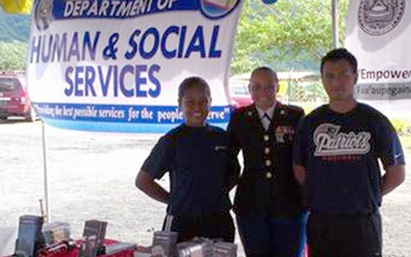 Three individuals are standing behind a U.S. Marine Corps Recruiting table at what appears to be a community outreach or job fair event. The table is covered with a red Marine Corps tablecloth and displays various recruitment materials and brochures. Behind them are banners for the Department of Human &amp; Social Services and an Employment America banner.