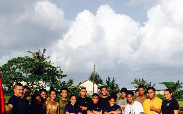 A group of approximately 15-18 individuals poses outdoors on a grass field, with a red USMC (United States Marine Corps) flag visible on the left side. The setting appears to be a tropical or subtropical location based on the palm trees, lush vegetation, and cumulus cloud formations. The group appears to be participating in a Marine Corps recruiting or fitness event.