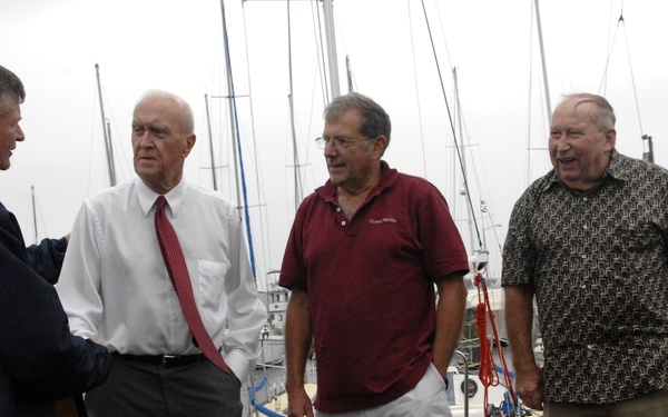 Four men are standing on a wooden dock at a marina, engaged in conversation. Two of the men appear to be shaking hands, suggesting a formal or semi-formal greeting or introduction. Multiple sailboats are moored in the background under an overcast sky.