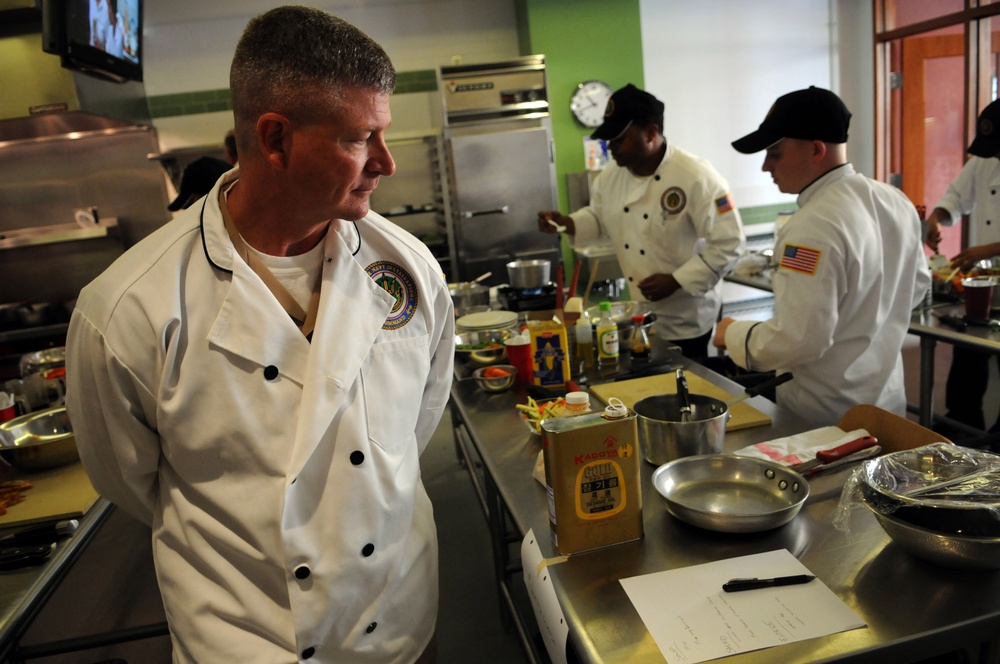 A professional kitchen scene showing multiple chefs in white chef coats working at stainless steel prep stations. The chefs appear to be participating in a culinary competition or training exercise, with various ingredients and cooking equipment visible on the counters. At least two of the chefs wear American flag patches on their sleeves, suggesting a military or government affiliation.