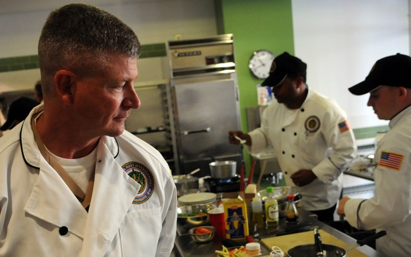 A professional kitchen scene showing multiple chefs in white chef coats working at stainless steel prep stations. The chefs appear to be participating in a culinary competition or training exercise, with various ingredients and cooking equipment visible on the counters. At least two of the chefs wear American flag patches on their sleeves, suggesting a military or government affiliation.