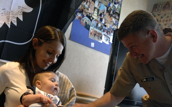 A military officer in tan/khaki Navy uniform is interacting with a woman holding an infant, likely in a hospital or medical facility room. The officer is leaning forward and pointing at or touching the baby while smiling. The setting appears to be a patient room with personal items and photos on the walls.