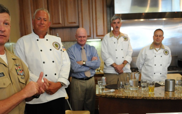 A U.S. Navy officer in khaki dress uniform addresses a group in what appears to be a culinary competition or food tasting event. Several individuals in white chef coats stand behind a kitchen counter, while plated dishes are displayed on a wooden table in the foreground. Name placards on the table indicate regional Navy districts including 'Northwest Region,' 'Naval District Washington,' and 'Hawaii.'