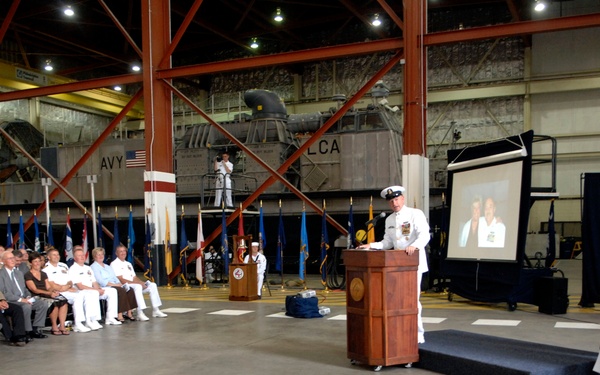 A formal U.S. Navy military ceremony, likely a change of command or retirement, taking place inside a large naval hangar facility. A naval officer in dress whites is speaking at a wooden podium before an audience of military personnel and civilians. A projection screen displaying what appears to be a photo is visible in the background alongside multiple flags.