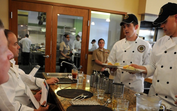 A culinary training or competition event appears to be taking place in a professional kitchen setting. Two chefs wearing white chef coats with embroidered patches and black baseball caps are presenting a plated dish to seated judges or evaluators. The setting includes a granite counter with place settings, water pitchers, and score sheets.