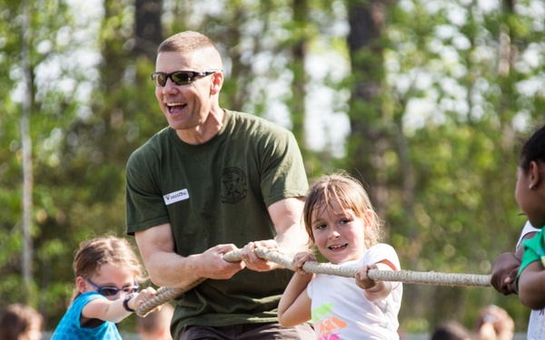 An adult male wearing a green military-style t-shirt and a visitor badge participates in a tug-of-war game with a group of children at an outdoor event. The scene appears to be a school or community field day activity, with multiple children pulling a thick rope together. The atmosphere is energetic and joyful, with the adult laughing and children showing focused determination.