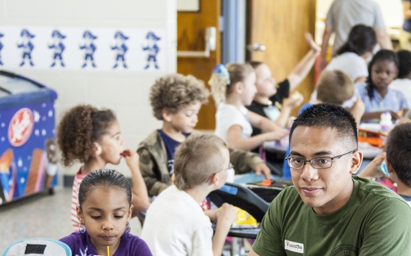 The image shows a school cafeteria or lunchroom setting where adults in military-style green t-shirts are seated alongside young children during a meal. The scene appears to be a special event, possibly a 'Lunch with a Loved One' or military family engagement day at an elementary school. Multiple tables are filled with children and adults sharing food and conversation in a lively, colorful environment.