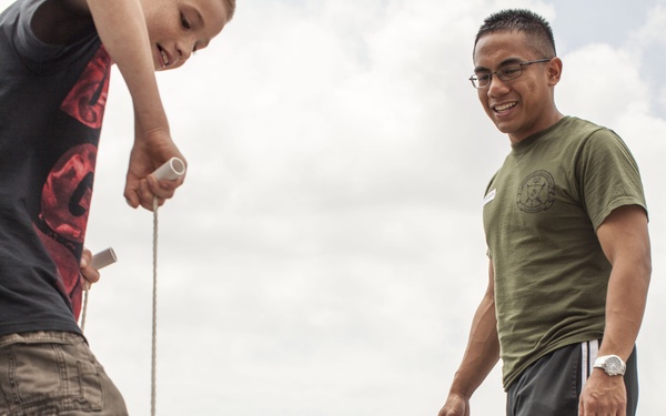 An outdoor event, likely a military family day or community activity, shows a young boy using a stick or rod near a pair of worn military combat boots placed on the ground. An adult male wearing a military-style olive green t-shirt with a unit insignia, black Nike track pants, and a watch watches with a smile. Multiple children and adults are visible in the background engaged in various activities on a paved surface.