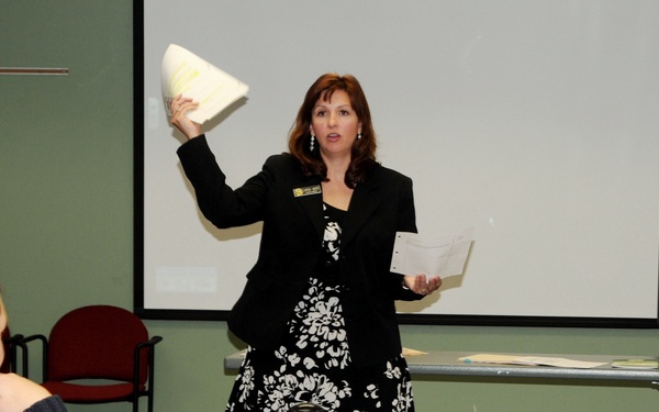 A woman in a black and white floral dress with a black blazer is presenting or leading a workshop/training session in a conference or classroom setting. She is holding up what appears to be a document or material in one hand while holding papers in the other. Two attendees are seated at a table in the foreground, with various materials, notebooks, and beverages on the table.