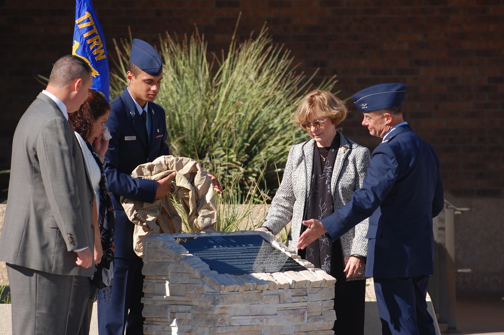 A formal military dedication or unveiling ceremony is taking place outdoors. Several individuals, including uniformed Air Force personnel and civilians, are gathered around a stone monument with a dark commemorative plaque being unveiled. The setting appears to be at a military installation or institutional building.