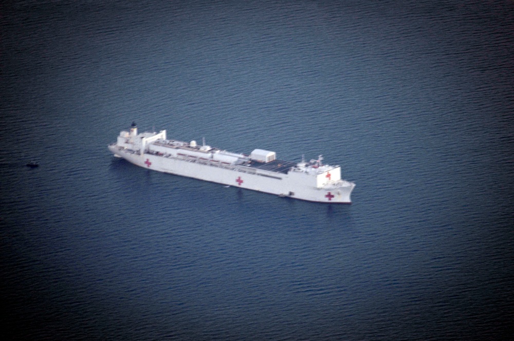 An aerial photograph of a large white hospital ship at sea, identifiable by the prominent red cross markings on its hull. The vessel appears to be stationary or moving slowly in calm, dark blue open water. The ship's all-white paint scheme and multiple red crosses are consistent with a military or international humanitarian hospital ship.