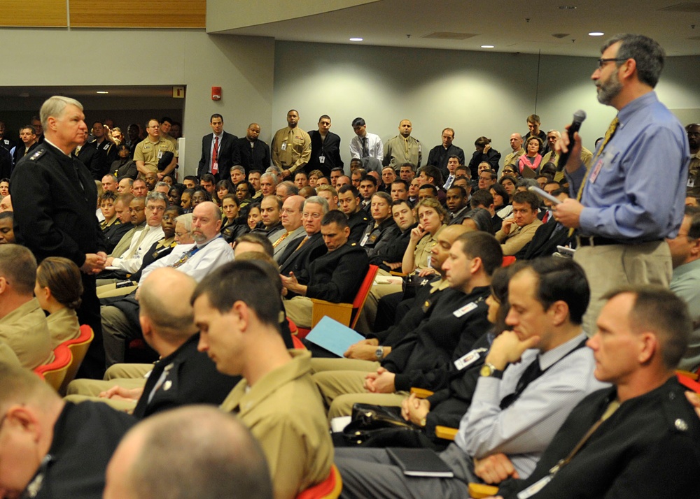 A large auditorium or conference hall filled with military personnel in various uniforms, along with some civilians. A civilian man in a blue shirt is standing and speaking into a microphone on the right side, while a uniformed officer stands at the front left. The audience appears to be attending a formal briefing, town hall, or all-hands meeting.