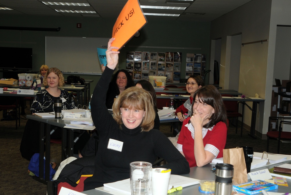 A group of women are seated at tables in what appears to be a classroom or workshop setting. A woman named 'Kacey' (visible on her name tag) is enthusiastically holding up an orange sign that reads 'PICK US!' while smiling at the camera. The setting suggests a training session, seminar, or team-building activity.