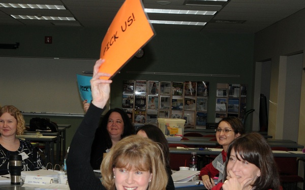 A group of women are seated at tables in what appears to be a classroom or workshop setting. A woman named 'Kacey' (visible on her name tag) is enthusiastically holding up an orange sign that reads 'PICK US!' while smiling at the camera. The setting suggests a training session, seminar, or team-building activity.