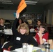A group of women are seated at tables in what appears to be a classroom or workshop setting. A woman named 'Kacey' (visible on her name tag) is enthusiastically holding up an orange sign that reads 'PICK US!' while smiling at the camera. The setting suggests a training session, seminar, or team-building activity.