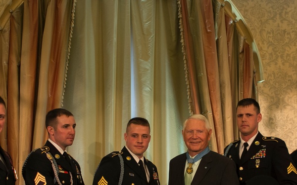 A formal military ceremony photograph showing five U.S. Army soldiers in dress uniforms posing with an elderly civilian man wearing a Medal of Honor around his neck. The group is standing in an ornate indoor venue with American flags and decorative draping in the background. This appears to be an official recognition or commemorative event.