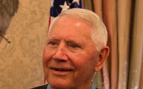 An elderly man wearing the United States Medal of Honor around his neck is engaged in conversation with another person whose back is to the camera. The setting appears to be a formal indoor venue with decorative wallpaper, curtains, and an American flag visible in the background. The man appears to be a Medal of Honor recipient at what looks like an official or ceremonial event.