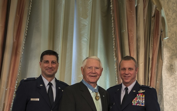Three men pose for a formal photograph in what appears to be a banquet or ceremonial hall. The central figure is an elderly civilian wearing a Medal of Honor around his neck, flanked by two U.S. Air Force officers in dress uniforms. The setting and attire suggest a formal military recognition or awards ceremony.