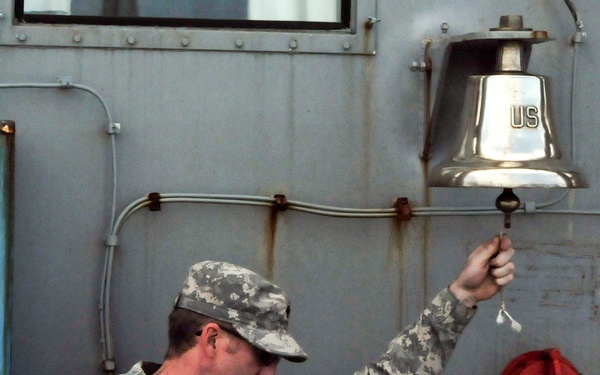 A U.S. Army soldier in ACU (Army Combat Uniform) digital camouflage is ringing a ship's bell marked 'US' aboard the USAV Matamoros (LCU-2026), a Landing Craft Utility vessel. The soldier's uniform displays an American flag patch and what appears to be a 3rd Infantry Division patch on the left sleeve. A display placard in the foreground provides information about the vessel and the LCU-2000 series.
