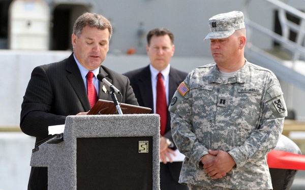 An outdoor press conference or official ceremony taking place at what appears to be a waterfront or dock facility. A civilian in a black suit and red tie is speaking at a podium, flanked by a U.S. Army soldier in ACU (Army Combat Uniform) and another civilian in a dark suit. The setting and participants suggest an official government or military-related announcement.