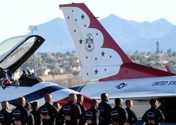 A group of approximately 12-13 United States Air Force Thunderbirds personnel stand in formation in front of an F-16 Fighting Falcon jet bearing the iconic Thunderbirds livery. The personnel are dressed in matching black uniforms and are holding helmets with the Thunderbirds logo. The scene appears to be at a desert airbase, likely in the southwestern United States, with mountains and suburban development visible in the background.