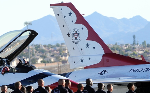 A group of approximately 12-13 United States Air Force Thunderbirds personnel stand in formation in front of an F-16 Fighting Falcon jet bearing the iconic Thunderbirds livery. The personnel are dressed in matching black uniforms and are holding helmets with the Thunderbirds logo. The scene appears to be at a desert airbase, likely in the southwestern United States, with mountains and suburban development visible in the background.