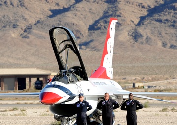 Three military personnel in flight suits stand in front of a U.S. Air Force Thunderbirds F-16 Fighting Falcon on a tarmac. The aircraft displays the distinctive red, white, and blue Thunderbirds livery with the canopy open. The scene appears to be at a desert air base, likely Nellis Air Force Base in Nevada, with rocky mountains visible in the background.