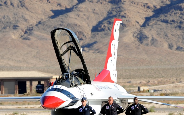 Three military personnel in flight suits stand in front of a U.S. Air Force Thunderbirds F-16 Fighting Falcon on a tarmac. The aircraft displays the distinctive red, white, and blue Thunderbirds livery with the canopy open. The scene appears to be at a desert air base, likely Nellis Air Force Base in Nevada, with rocky mountains visible in the background.