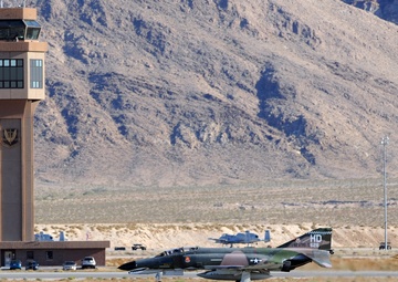 A camouflage-painted F-4 Phantom II jet aircraft is landing on a runway at a desert military airbase, deploying a yellow drag parachute for deceleration. The aircraft bears the tail code 'HD 826' and US Air Force markings, with a distinctive Southeast Asia-era green and brown camouflage scheme. A control tower with an eagle emblem, arid mountain terrain, and sparse desert vegetation are visible in the background.