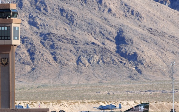 A camouflage-painted F-4 Phantom II jet aircraft is landing on a runway at a desert military airbase, deploying a yellow drag parachute for deceleration. The aircraft bears the tail code 'HD 826' and US Air Force markings, with a distinctive Southeast Asia-era green and brown camouflage scheme. A control tower with an eagle emblem, arid mountain terrain, and sparse desert vegetation are visible in the background.