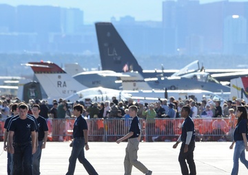 A group of young people wearing matching navy blue shirts walk across an airfield tarmac at what appears to be a military air show. Multiple military aircraft are visible in the background, along with a large crowd of spectators behind orange safety barriers. A city skyline is visible in the distant background, suggesting this is near a major urban area.