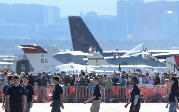 A group of young people wearing matching navy blue shirts walk across an airfield tarmac at what appears to be a military air show. Multiple military aircraft are visible in the background, along with a large crowd of spectators behind orange safety barriers. A city skyline is visible in the distant background, suggesting this is near a major urban area.