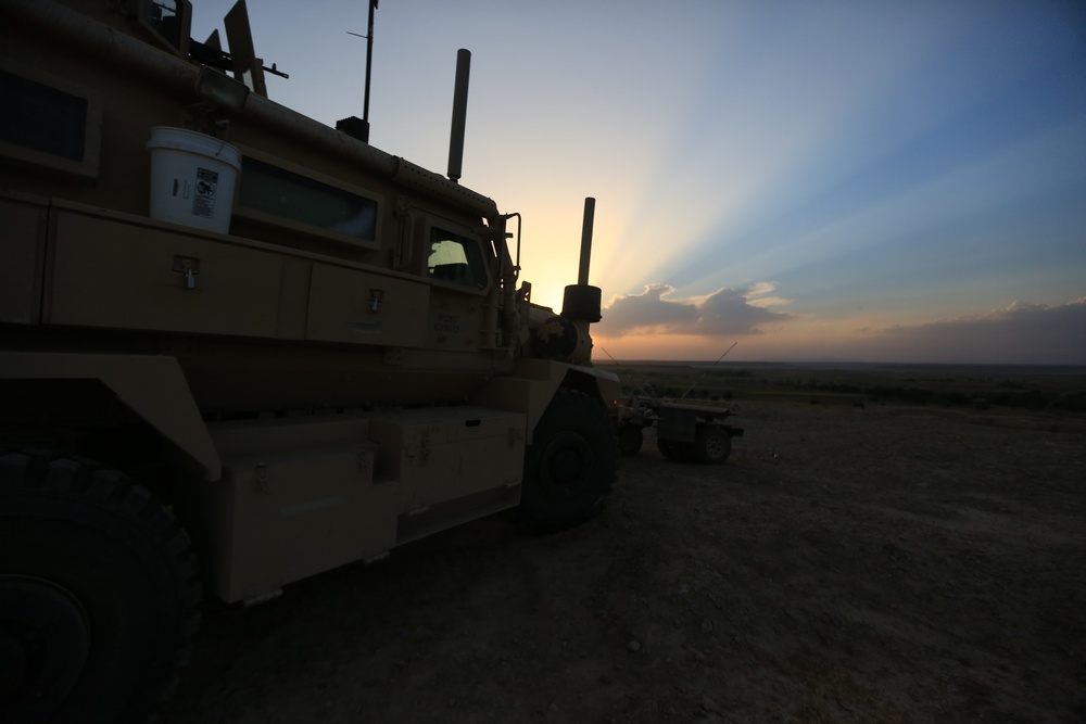 A tan/desert-camouflaged military armored vehicle, likely an MRAP (Mine-Resistant Ambush Protected) vehicle, is parked on flat, arid terrain at sunset or sunrise. Crepuscular rays radiate from behind clouds on the horizon, creating a dramatic sky. A second military vehicle is partially visible in the background.