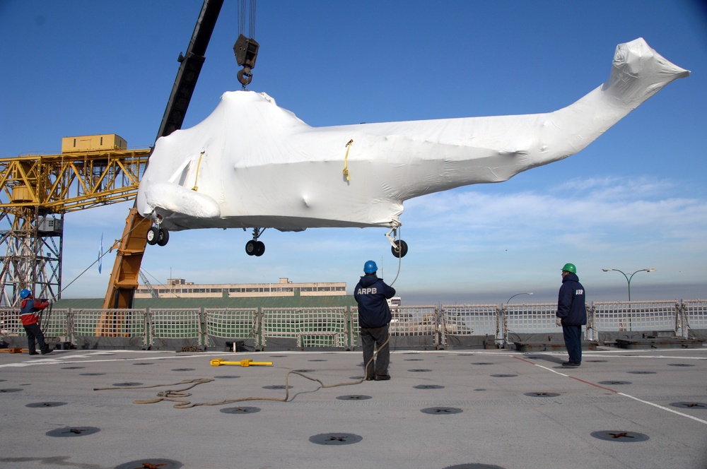 A shrink-wrapped helicopter is being lifted by a yellow crane onto what appears to be the deck of a naval vessel or pier. Three workers wearing blue and green hard hats and jackets marked 'ARPB' are guiding the operation. The helicopter's landing gear is visible beneath the white protective wrapping, and the scene takes place near a waterfront with buildings visible in the background.