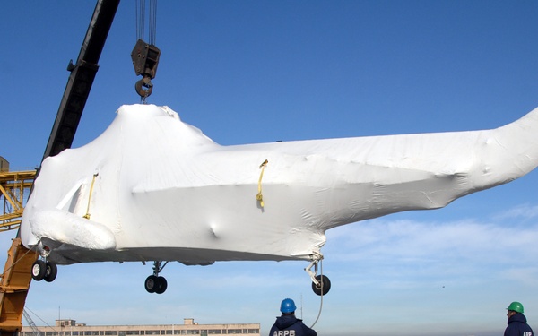 A shrink-wrapped helicopter is being lifted by a yellow crane onto what appears to be the deck of a naval vessel or pier. Three workers wearing blue and green hard hats and jackets marked 'ARPB' are guiding the operation. The helicopter's landing gear is visible beneath the white protective wrapping, and the scene takes place near a waterfront with buildings visible in the background.