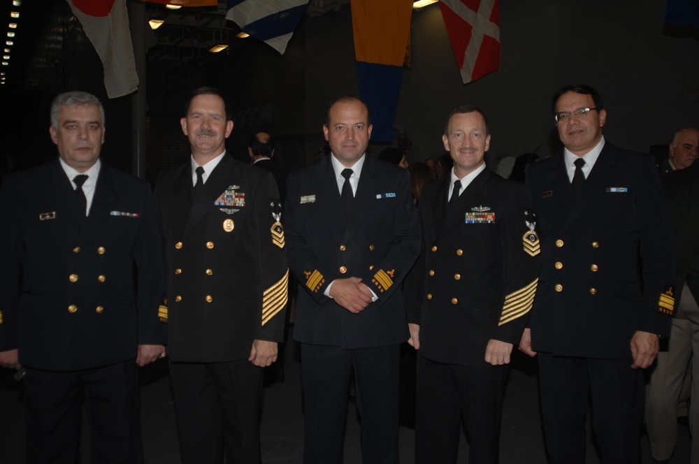 Five military personnel in dark naval dress uniforms are posed together for a group photograph at what appears to be a formal event or ceremony. The individuals display various ranks indicated by gold sleeve stripes, medals, and insignia on their uniforms. Multiple national flags are visible hanging in the background, suggesting an international or multinational military gathering.