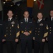 Five military personnel in dark naval dress uniforms are posed together for a group photograph at what appears to be a formal event or ceremony. The individuals display various ranks indicated by gold sleeve stripes, medals, and insignia on their uniforms. Multiple national flags are visible hanging in the background, suggesting an international or multinational military gathering.