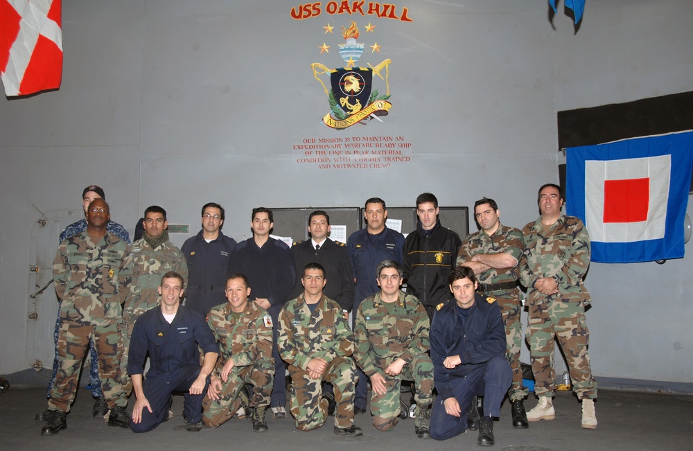 A group of approximately 15 military personnel pose for a formal group photograph aboard the USS Oak Hill, a U.S. Navy expeditionary warfare ship. The personnel are wearing a variety of military uniforms including camouflage, dress blues, and black jackets, suggesting a multinational or multi-branch gathering. The group is arranged in two rows — standing and kneeling — in front of the ship's official insignia and mission statement.
