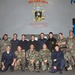 A group of approximately 15 military personnel pose for a formal group photograph aboard the USS Oak Hill, a U.S. Navy expeditionary warfare ship. The personnel are wearing a variety of military uniforms including camouflage, dress blues, and black jackets, suggesting a multinational or multi-branch gathering. The group is arranged in two rows — standing and kneeling — in front of the ship's official insignia and mission statement.