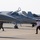A military flight line showing multiple jet fighter aircraft parked on a tarmac apron. Personnel in various uniforms are walking between the aircraft, and at least one pilot is visible in a cockpit. The aircraft include what appears to be an F-16 in the foreground and multiple F/A-18 Hornets bearing VMFA-314 markings behind it.