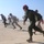 Military personnel are running across a tarmac or airfield in what appears to be a training exercise or scramble drill. The group includes individuals in flight suits and camouflage uniforms, with one person wearing a distinctive red flight helmet. The setting appears to be a desert airbase based on the arid, flat terrain visible in the background.