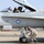 A U.S. Marine Corps F/A-18 Hornet fighter jet is on a flight line with a pilot seated in the cockpit and a ground crew member inspecting or working on the nose landing gear. The aircraft bears the squadron designation 'VMFA' and is assigned to Lt. Col. B.D. Wild with callsign 'HOG'. The ground crew member, identified by markings as Sgt. B.C. McDowell from Chicago, IL, is wearing desert camouflage utilities and hearing protection.