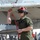 A military aviation technician or ordnanceman in olive green flight suit is performing a pre-flight or post-flight inspection on an air-to-air missile mounted under the wing of a military jet aircraft. The individual is wearing a red flight deck helmet with visor and hearing protection, and is holding a red safety pin lanyard. The setting appears to be a flight deck or airfield with other personnel visible in the background.