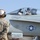 A military jet aircraft, likely an F/A-18 Hornet, is shown on what appears to be a flight deck or airfield. The aircraft is assigned to LTCOL B.D. WILD with callsign 'HOG', and the crew chief placard reads SGT B.C. McDOWELL from Chicago, IL. A ground crew member wearing a 'Power Line' helmet is facing the aircraft while the pilot waves from the open cockpit.