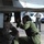 Two military personnel are crouching beneath an aircraft on a flight line, closely inspecting or working on an undercarriage component. One individual is wearing Marine Corps camouflage utilities while the other wears an olive green flight suit, both equipped with communication headsets. In the background, several personnel in Air Force uniforms observe from a distance near parked vehicles.