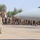 A military ceremony is taking place on an airfield tarmac, with a uniformed soldier in the foreground raising both hands toward a large formation of troops lined up alongside a large military aircraft. A Jordanian flag is visible among the assembled personnel, suggesting this is a joint military event or exercise in Jordan. The scene appears to be a formal greeting, inspection, or ceremonial arrival/departure.