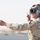 A military personnel wearing desert digital camouflage uniform is giving hand signals, likely directing aircraft or vehicles on a flight line or airfield. The individual is equipped with a flight deck helmet, protective goggles, and a communications headset with boom microphone. The arid, hazy background suggests a desert environment.