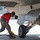 A military aircraft maintainer in a red shirt and camouflage pants is working beneath a military jet aircraft on a flight line or tarmac. The individual appears to be placing or removing wheel chocks near the nose or main landing gear of the aircraft. The scene takes place outdoors in daylight at what appears to be a military airfield.