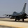 Two military fighter jets are taxiing on a runway at what appears to be a desert airbase. The foreground aircraft is an F-16 Fighting Falcon bearing tail code 'WW' and serial number 891, while a second aircraft, likely an F/A-18 Hornet, is visible in the background. The arid, flat terrain and hazy sky suggest a desert environment.