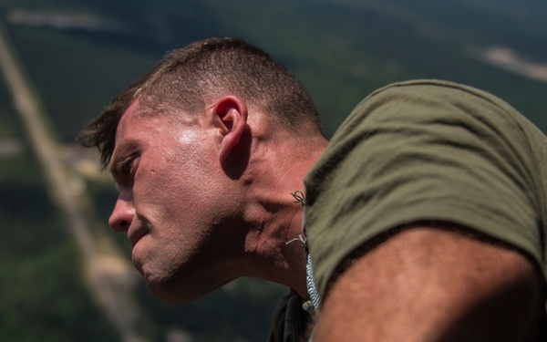 A male individual wearing an olive green t-shirt is leaning out from what appears to be an aircraft at significant altitude, looking downward toward a landscape below. The ground visible in the background shows green terrain with roads or paths cutting through it. The person appears to be sweating and is equipped with some form of harness or gear.